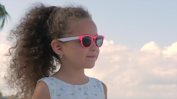 Close-up Portrait of a Beautiful Little Girl in Pink Glasses, Cute Smiling, Looking at the Blue Sky alt