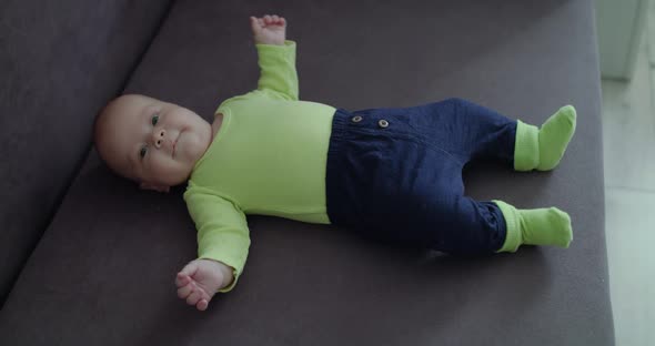Surprised Baby Boy Lying on Sofa with Open Arms and Looking Into Camera in Home alt
