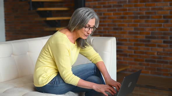 Beautiful Positive Middleaged Woman Sitting on the White Couch and Using Laptop alt