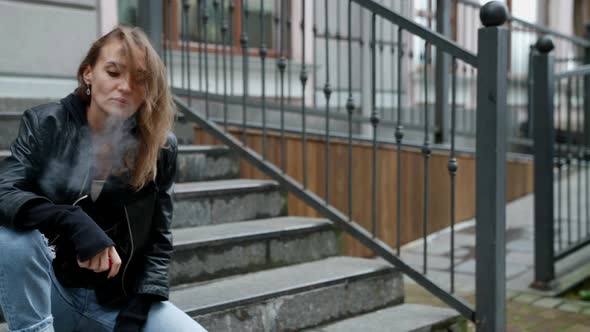 a Sad Blonde in Ripped Jeans and a Black Jacket is Sitting on the Stairs at Home alt