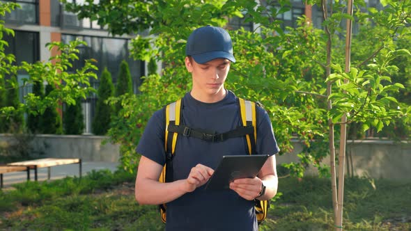 Zoom in Serious Food Delivery Man Courier with Thermal Backpack Uses Tablet alt