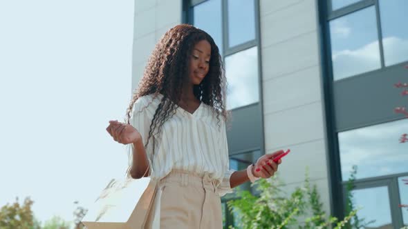 Excited Black Woman Customer Walking on Street Holding Shopping Bags and Phone alt