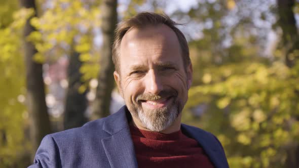 A Middleaged Caucasian Man Smiles at the Camera As He Sits on a Bench in a Quaint Town in Fall