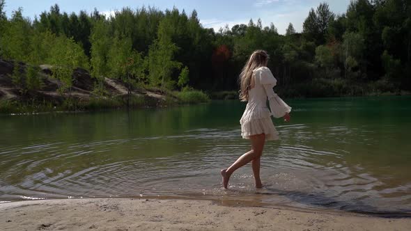 One Young Woman in a White Dress Is Walking and Wetting Her Feet on the Shore of a Wild Forest Lake alt