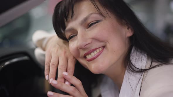 Closeup Face of Excited Happy Beautiful Woman Posing in Car on Driver's Seat in Dealership alt