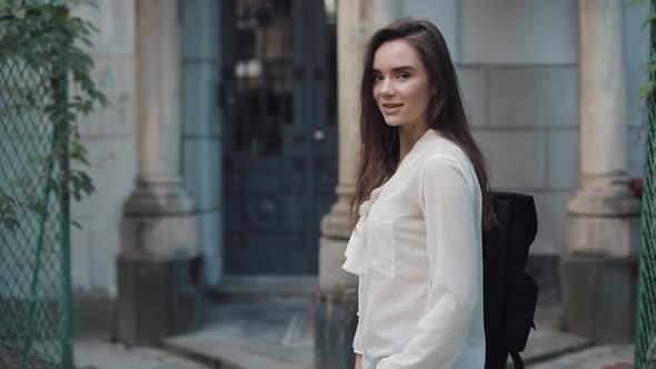 Portrait of Young Charming Brunette Student Girl with a Bag Standing Sideways and Looking To Camera alt