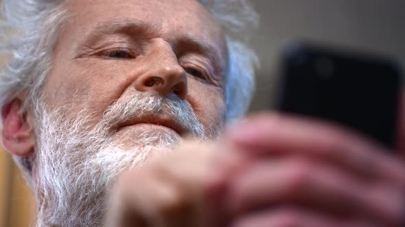 A Grayhaired Old Man with a Beard Holds a Smartphone in His Hand Uses It alt