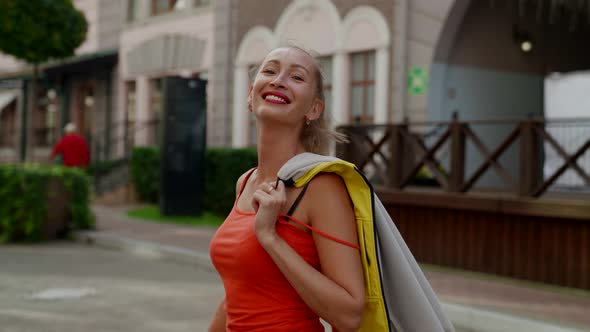 a Woman in Red Clothes Has Thrown Jacket Over Shoulder and is Walking Along a City Street Smiling alt