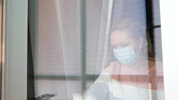 Sad Woman on Quarantine in Medical Mask Cleaning Window. People on Self Isolation alt