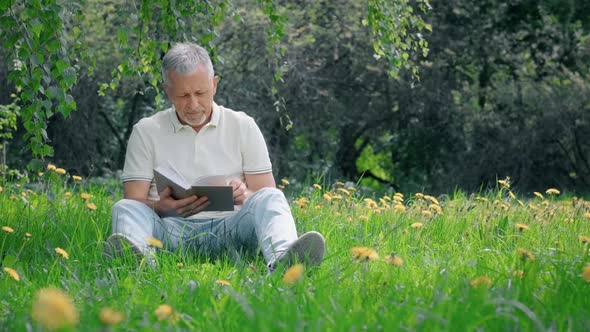 An Elderly Whitehaired Man with a Beard Sitting Alone on the Grass in a Nature Park with a Book