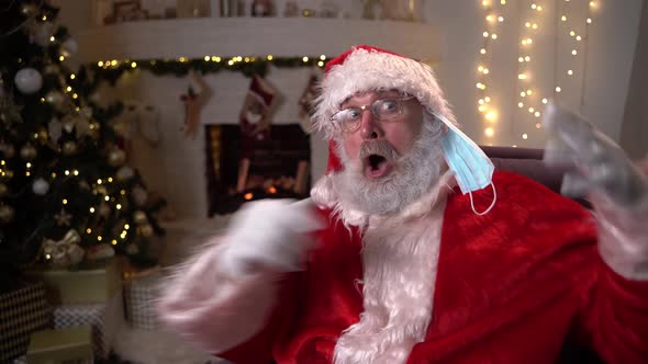 Funny Santa Claus Puts on a Protective Medical Mask While Sitting in a Chair By the Fireplace