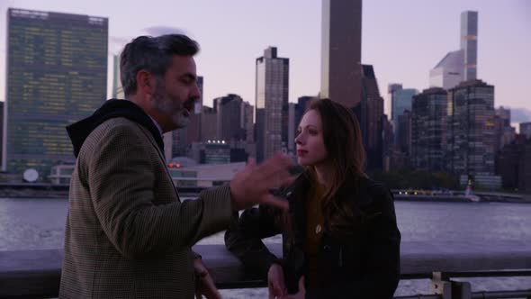 Couple in New York City stand by river talking with skyline in background