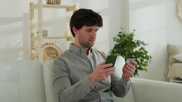 Man Sitting on the Sofa Holding Debit Card and Using Smartphone for Shopping at Home alt