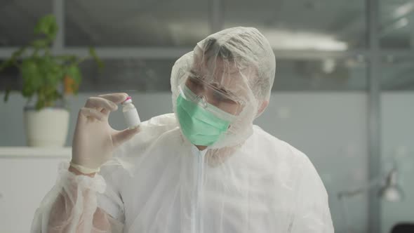 A Doctor in a Medical Protective Suit Holds a Bottle of Coronavirus Vaccine in His Hand and Examines alt
