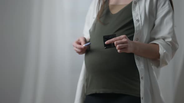 Pregnant Unrecognizable Caucasian Woman Holding Pregnancy Test and Condom Standing Indoors alt