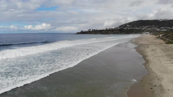 A drone flies over crashing ocean waves at the beach as surfers paddle out to surf. alt