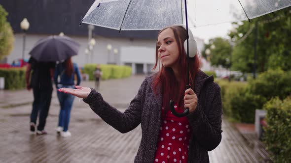 Young Beautiful Woman in Headphones Listening to Music Standing with Transparent Umbrella in Rain on alt