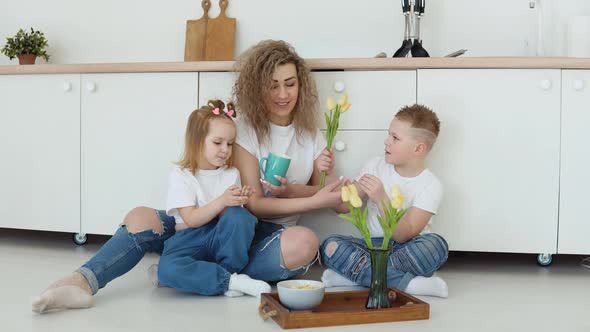 A Boy a Girl and a Mother Sit on the Floor in a Stylish White Modern Kitchen and Have Breakfast with alt