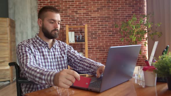 Man with Special Needs with Credit Card in His Hand is Sitting Wheelchair Front of Laptop Filling alt