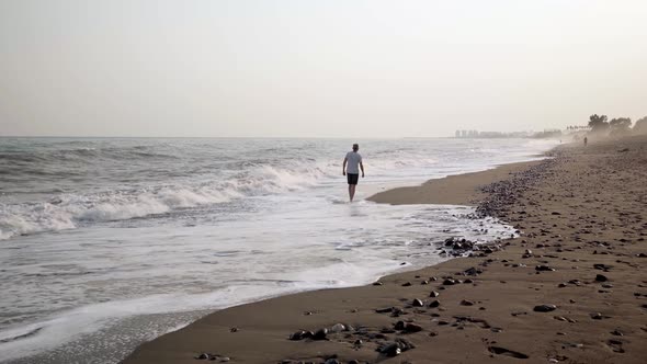 An Elderly Slender Man Walks Barefoot on a Sandy Beach in the Fastmoving Big Waves on a Summer