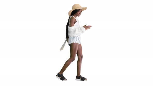 Pretty African American Woman in a Straw Hat Texting on Her Phone While Walking on White Background. alt