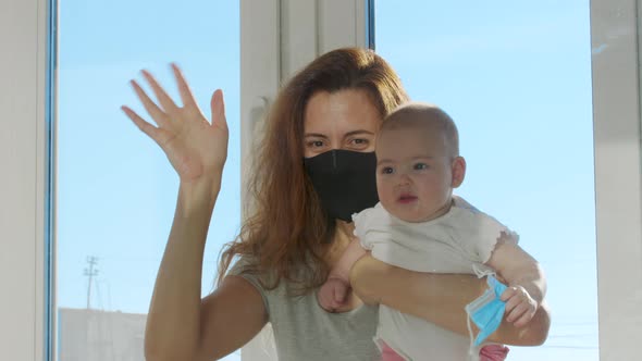 Mother in Medical Mask Communicated with Baby on Hands Through Glass Window alt