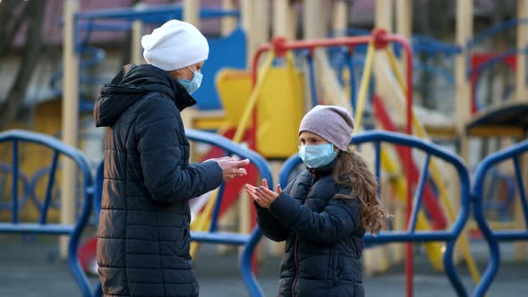 A girl in a medical mask. Her mother sprays disinfectant on her hands in the Playground. 
