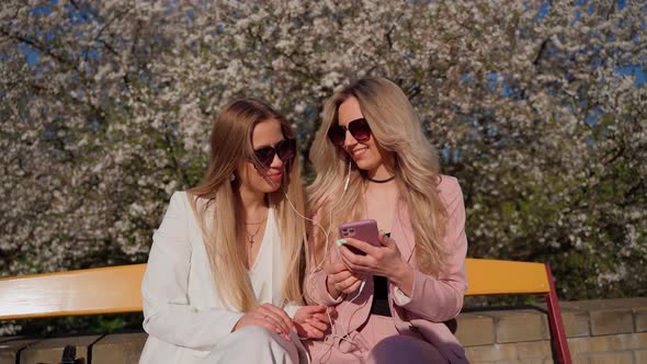 Two females persons looking at smartphone smile and laugh outside. Young adult women in sunglasses alt
