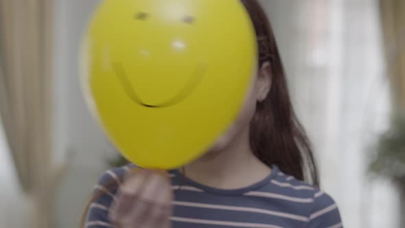 A Pretty Girl Hiding Behind Yellow Balloon with a Painted Happy Face on It Then Puts It Down alt