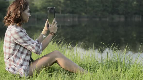 Young Woman in Nature Seated on Grass Take Photo with Tablet on Lake Shore in Sunny Summer Day