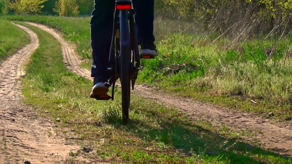 Young Guy Rides a Bicycle Along a Dusty Path in the Forest at Sunset alt