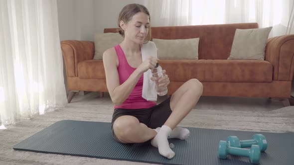 A Girl Drinks Water After a Workout at Home Sitting on a Mat Next to Dumbbells