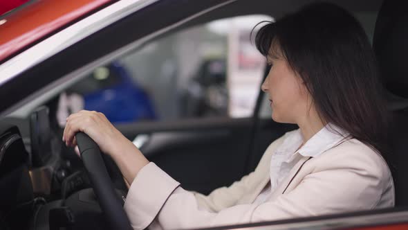 Side View of Confident Brunette Woman Sitting on Driver's Seat Choosing Vehicle in Car Dealership alt