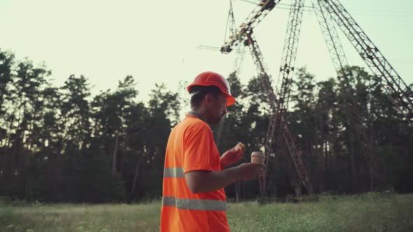 Hungry Worker in Overalls in Electric Power Business Eats Sandwich and Drinks Takeaway Coffee at alt