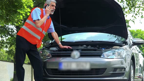 Senior Man Wears Warning Vest, Controls Engine of the Car and He Calls with Smartphone alt