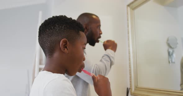 African american dad and son brushing teeth together looking in the mirror at home alt