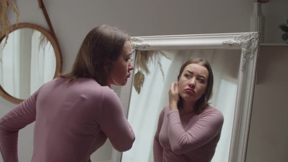 Displeased Charming Adult Woman Looking at Mirror Concerned About Wrinkles and Skin Aging alt