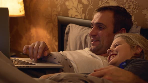 Footage of Middleaged Father with Light Bristle Lying in Bed with Laptop and His Small Blonde Child alt