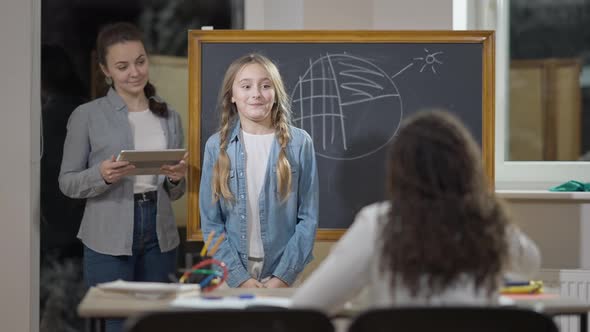 Portrait of Smart Cute Caucasian Schoolgirl Talking and Smiling Standing in Classroom with Teacher alt