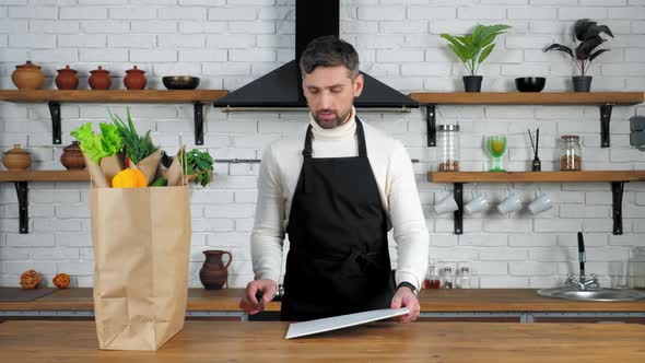 Man chef in black apron puts knife and board for cutting vegetables on kitchen alt