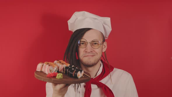 Young Man in Glasses Dressed As Chef Holding Wooden Board with Sushi Rolls alt