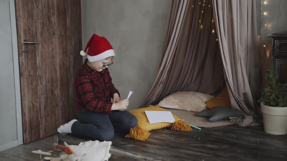 Caucasian Boy Wearing Glasses and a Santa Cap Puts a Letter in a Craft Envelope alt