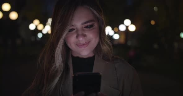 Cute Girl Uses Smartphone and Smiles Standing on Night Street in the City. Woman Messaging on Phone alt