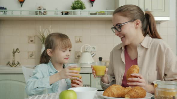 Young Woman and Little Girl Eating Croissants and Drinking Juice Talking at Breakfast alt
