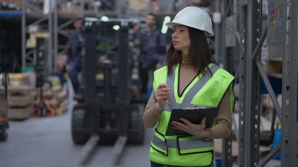 Concentrated Female Freight Agent in Hard Hat Standing in Warehouse Filling in Bill of Lading As Men alt