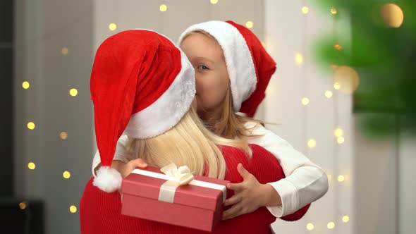 Happy Girl Hugs and Kisses Mother with Gift Box on Background of Christmas Tree