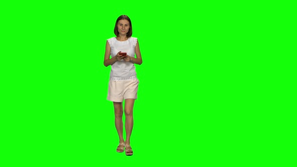 Young Woman Is Calmly Walking and Texting Message Vie Her Mobile Phone on Green Screen. Front View. alt