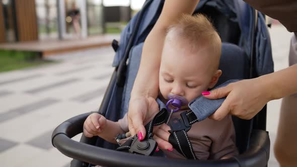 Mom Fastens the Baby in the Stroller for Safety on a Summer Day alt