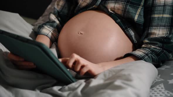Pregnant with tablet computer in bedroom showing and touching belly.