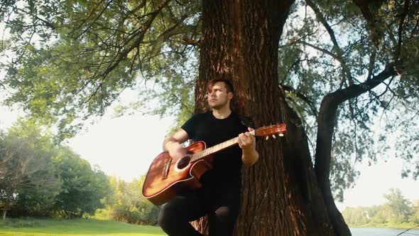A young guy in a black T-shirt plays an acoustic guitar leaning against a tree.